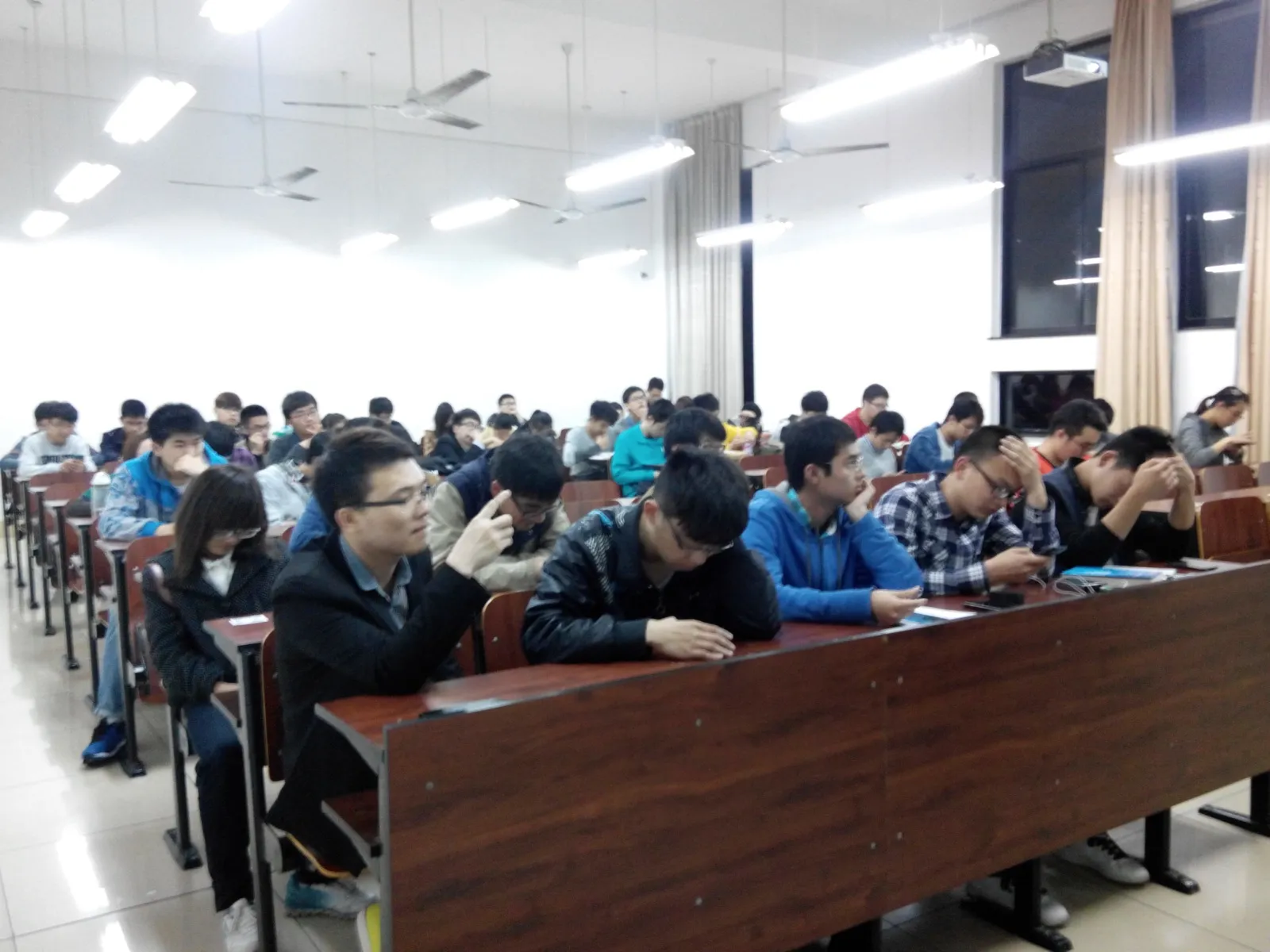 阶梯教室里坐满学生，前排几位撑着下巴或低头看手机，后排一直坐到窗边；右上角天花板上装着投影仪，室内日光灯全开