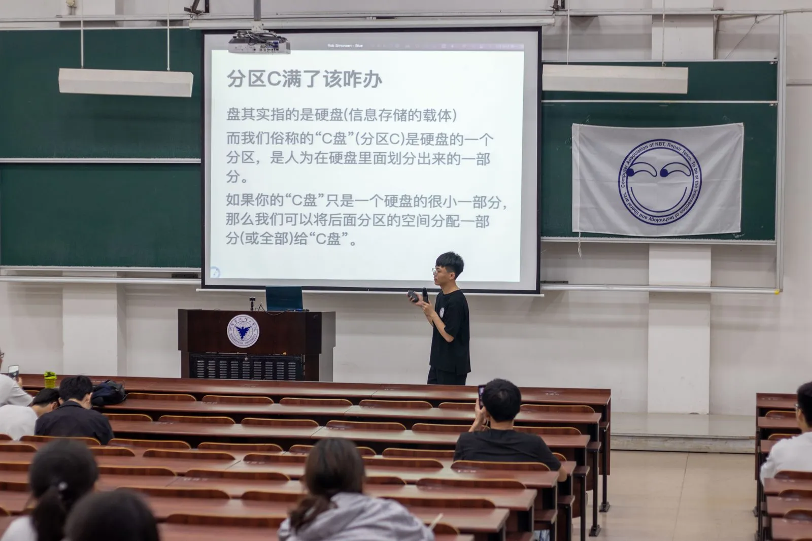 讲座投影上写着“分区C满了该咋办”,正解释“盘”指硬盘、而 C 盘只是硬盘上人为划出的一个分区;讲台右侧挂着协会队旗