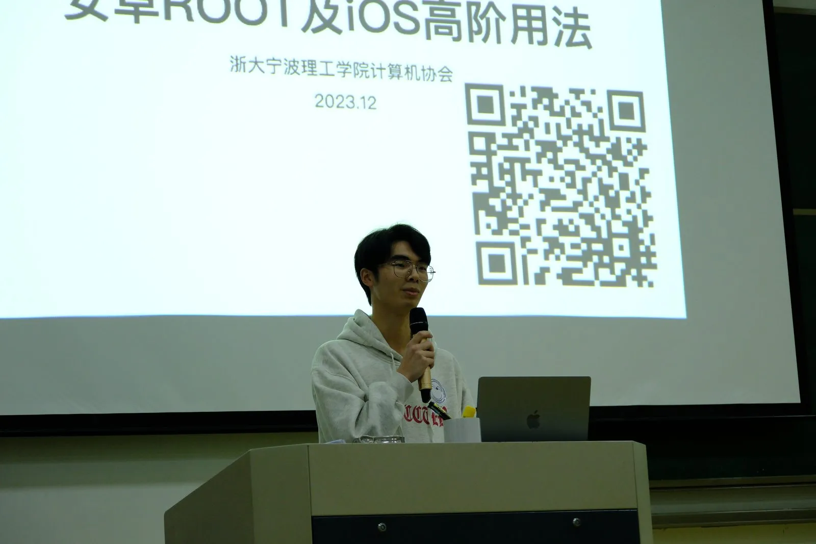 讲台上一名成员持话筒讲解,身后投影的标题页写着“安卓 ROOT 及 iOS 高阶用法/浙大宁波理工学院计算机协会/2023.12”,右侧有一个二维码