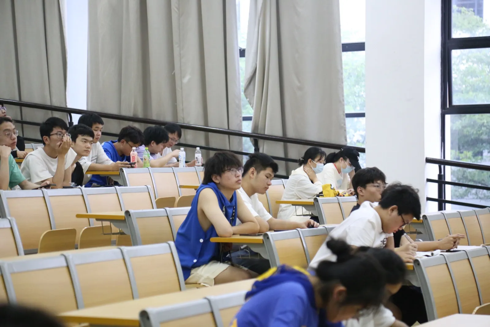 阶梯教室里学生分散坐在几排座位上，多数侧身朝向讲台方向，有人托腮听讲，有人低头做笔记，桌上放着饮料瓶；右侧是整面落地窗，窗外绿树