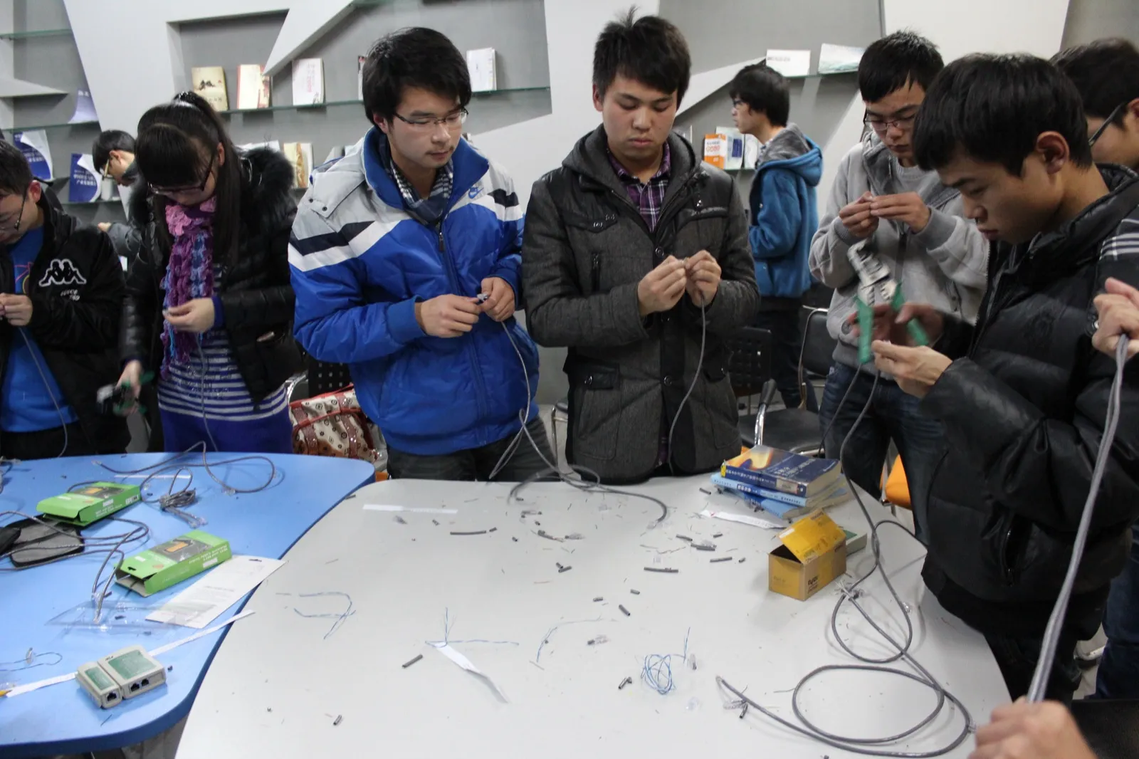 十余名学生穿着冬装围站在一张长桌四周，各自手里捏着一段网线在压接水晶头。桌上散落着剪下的线头与水晶头，还放着几把绿色压线钳、两台网线测试仪和一盒水晶头，背景是书架