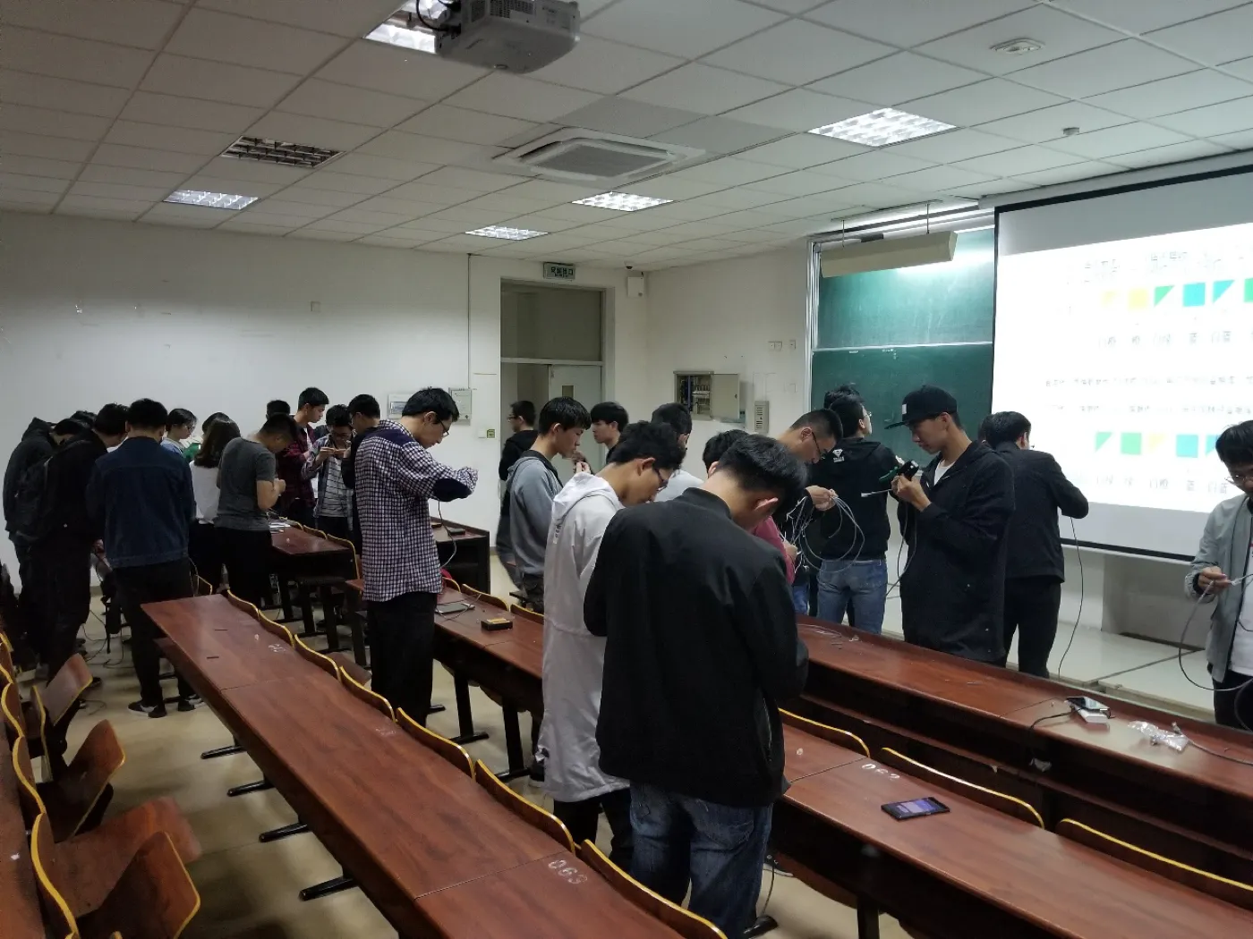 教室里两排长条课桌之间站满学生，各自低头摆弄手里的网线与工具，桌面上散着水晶头和线头，前方投影幕仍亮着