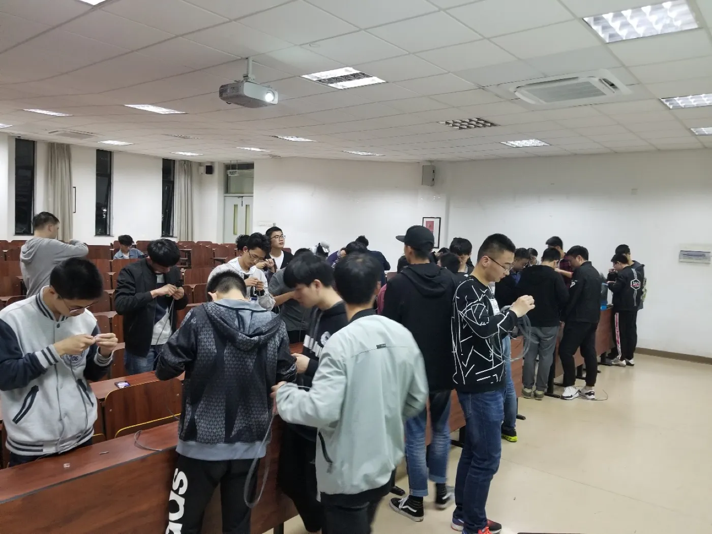 近处几名学生并排站着，手里各捏一段网线在理线序，旁边有人侧身指点；背景是亮着幻灯片的投影幕与其他围站的同学