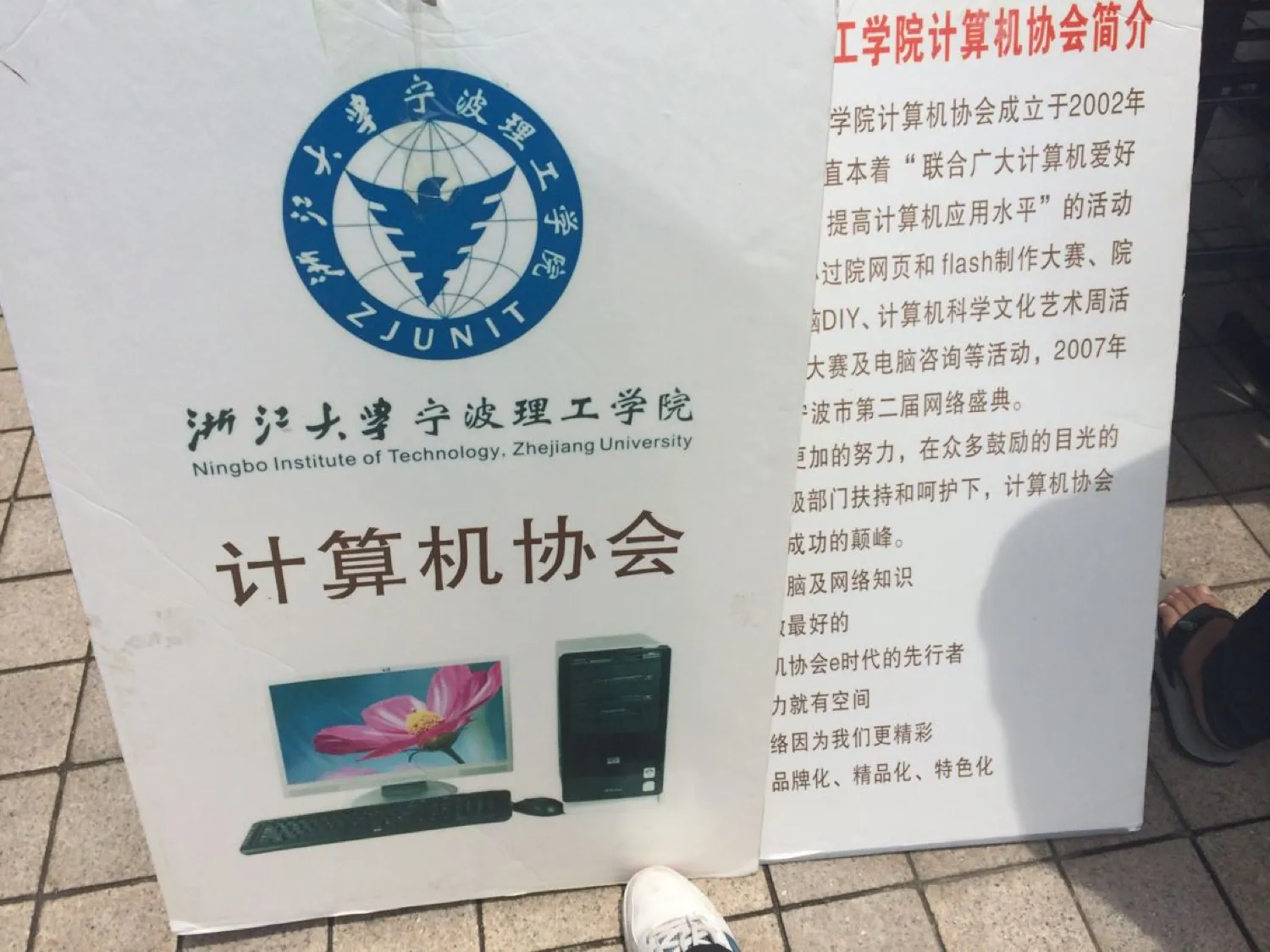 地砖上斜靠着两块白色 KT 板。左板上方是浙江大学宁波理工学院的蓝色校徽与中英文校名，下方大字“计算机协会”，再下面印着一台台式机与显示器的图片；右板红字标题“工学院计算机协会简介”，正文被左板遮去左半，仍可读出“……学院计算机协会成立于2002年”“……直本着‘联合广大计算机爱好……’”“……过院网页和 flash 制作大赛”“……脑DIY、计算机科学文化艺术周活……”“2007年……宁波市第二届网络盛典”，末行为“品牌化、精品化、特色化”