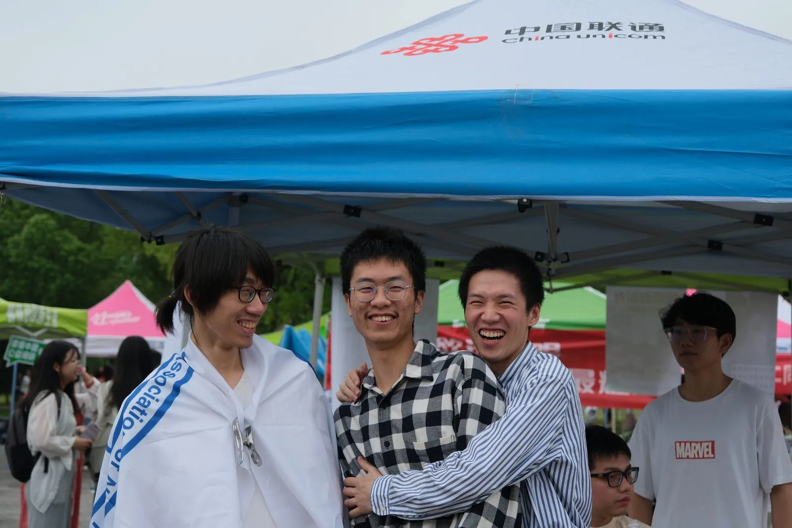 百团大战现场,三名成员在蓝白帐篷下搂着肩大笑,其中一人披着印有 Association 字样的绶带,背景是其他社团的粉色与绿色摊位