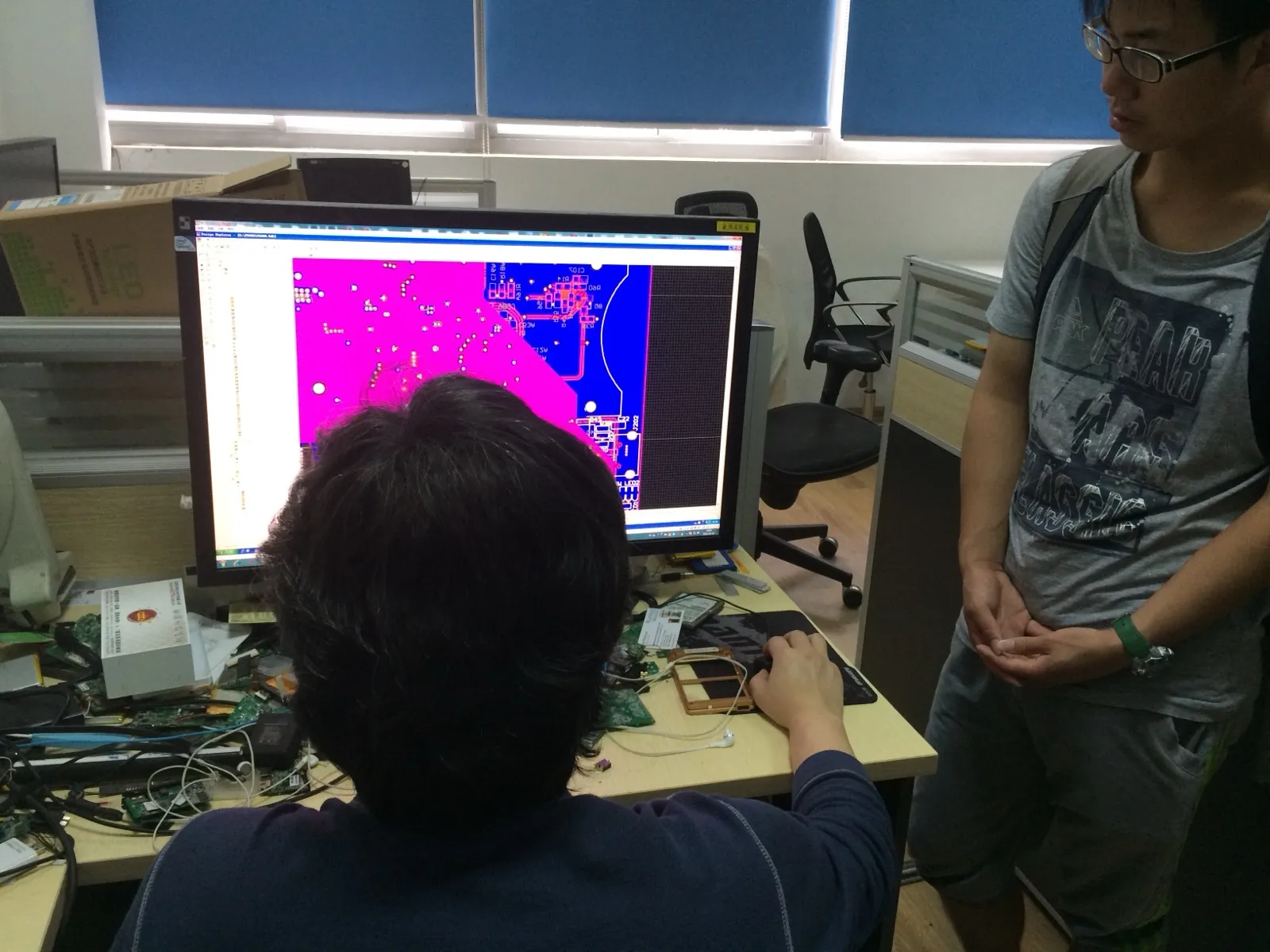 办公室工位上,一名工程师背对镜头坐着,面前的显示器上是 PCB 设计软件的布线视图,粉紫与蓝色的铜层叠在一起;桌面散放着多块拆下的电路板、螺丝刀与一块电池,右侧一名背着双肩包的学生站着看屏幕
