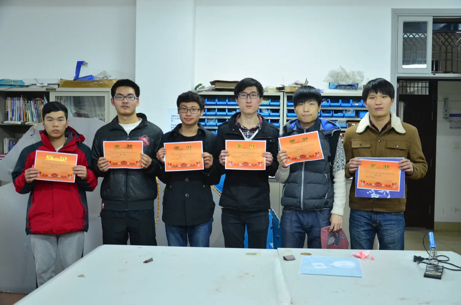 六名学生并排站着,每人双手举起一张橙色奖状,奖状上方印“奖状”二字,下方一行小字标着名次;背景是实验室的书架与一排排蓝色零件盒,桌上放着电烙铁与工具