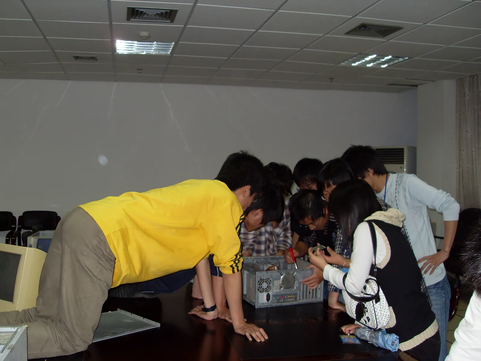会议室的深色长桌上放着一台侧板已卸下的台式机机箱，八九名学生围成半圈俯身向内看；一人手持红柄螺丝刀伸进机箱，另一人捧着一根内存条端详。左侧边缘是一台 CRT 显示器，卸下的侧板斜靠在桌上