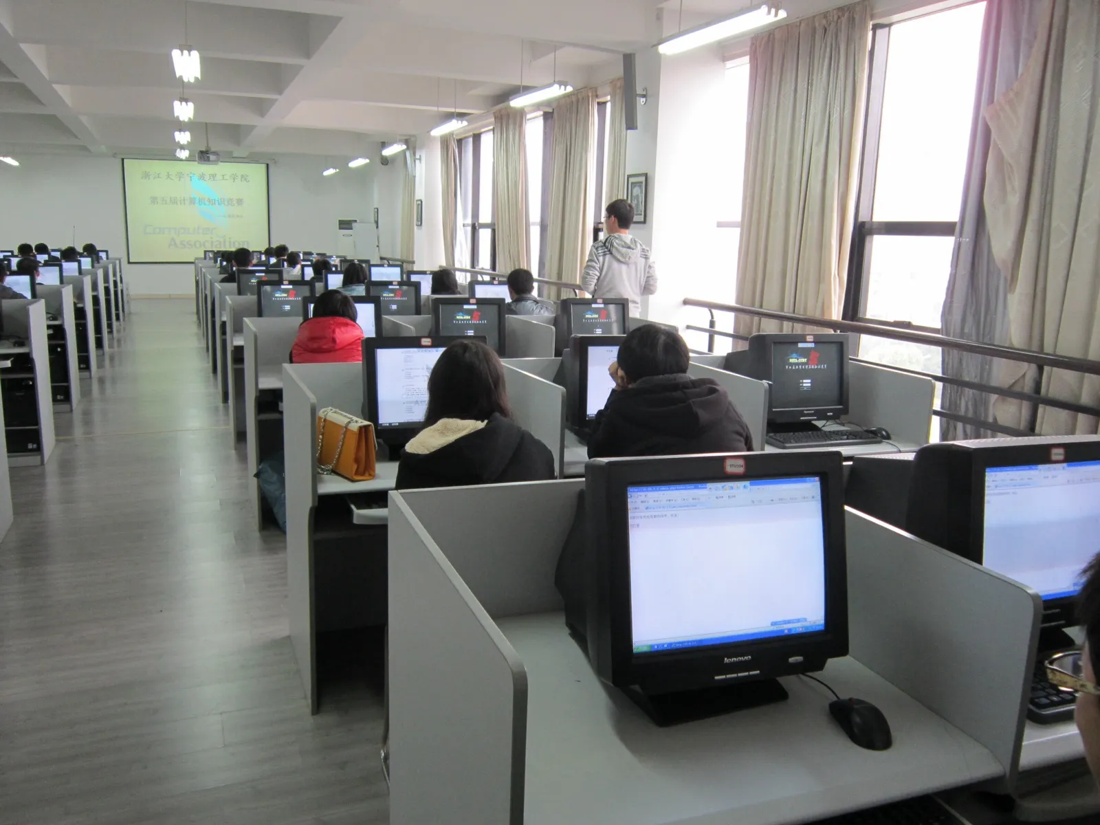 机房里带隔板的独立机位一排排铺开,学生分坐其间答题。左前方投影幕上写着“浙江大学宁波理工学院 第五届计算机知识竞赛”与 Computer Association 字样,多台显示器停在竞赛系统的登录页,一名工作人员站在窗边