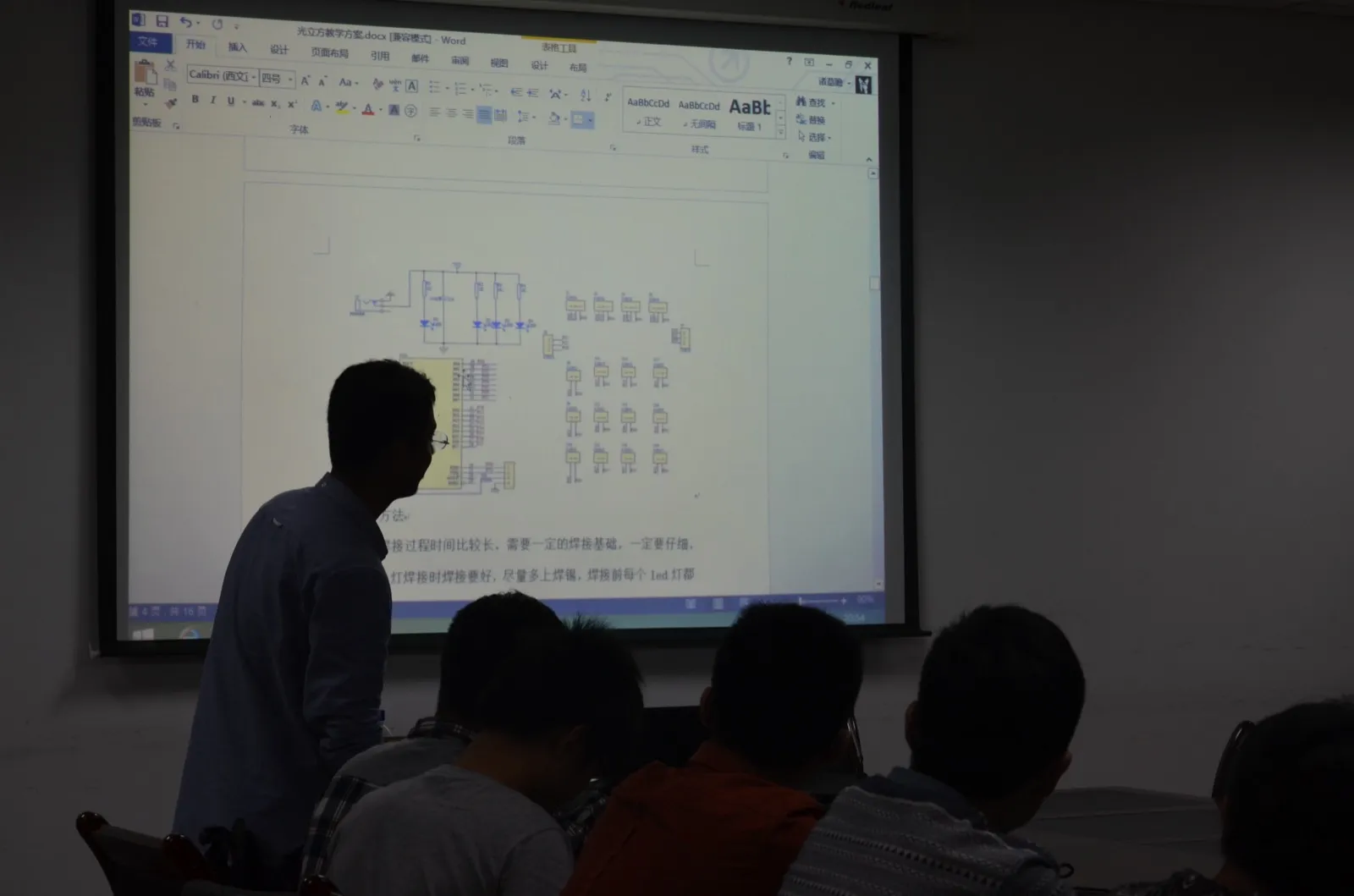 昏暗的教室里,投影幕上是一份名为“光立方教学方案.docx”的 Word 文档,页面上方为电路原理图,下方可见“焊接过程时间比较长,需要一定的焊接基础,一定要仔细”等文字;一名讲者背对镜头站在幕布前,前排几位学生仰头看着