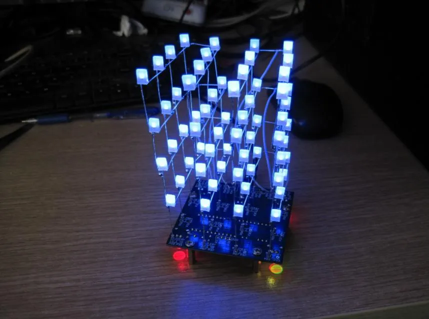 桌面上点亮的光立方:蓝色底板立起四层共 64 颗方形 LED,全部发蓝光,细导线在灯珠之间连成网格,底板边缘另有一红一黄两颗指示灯亮着,背景是键盘与鼠标