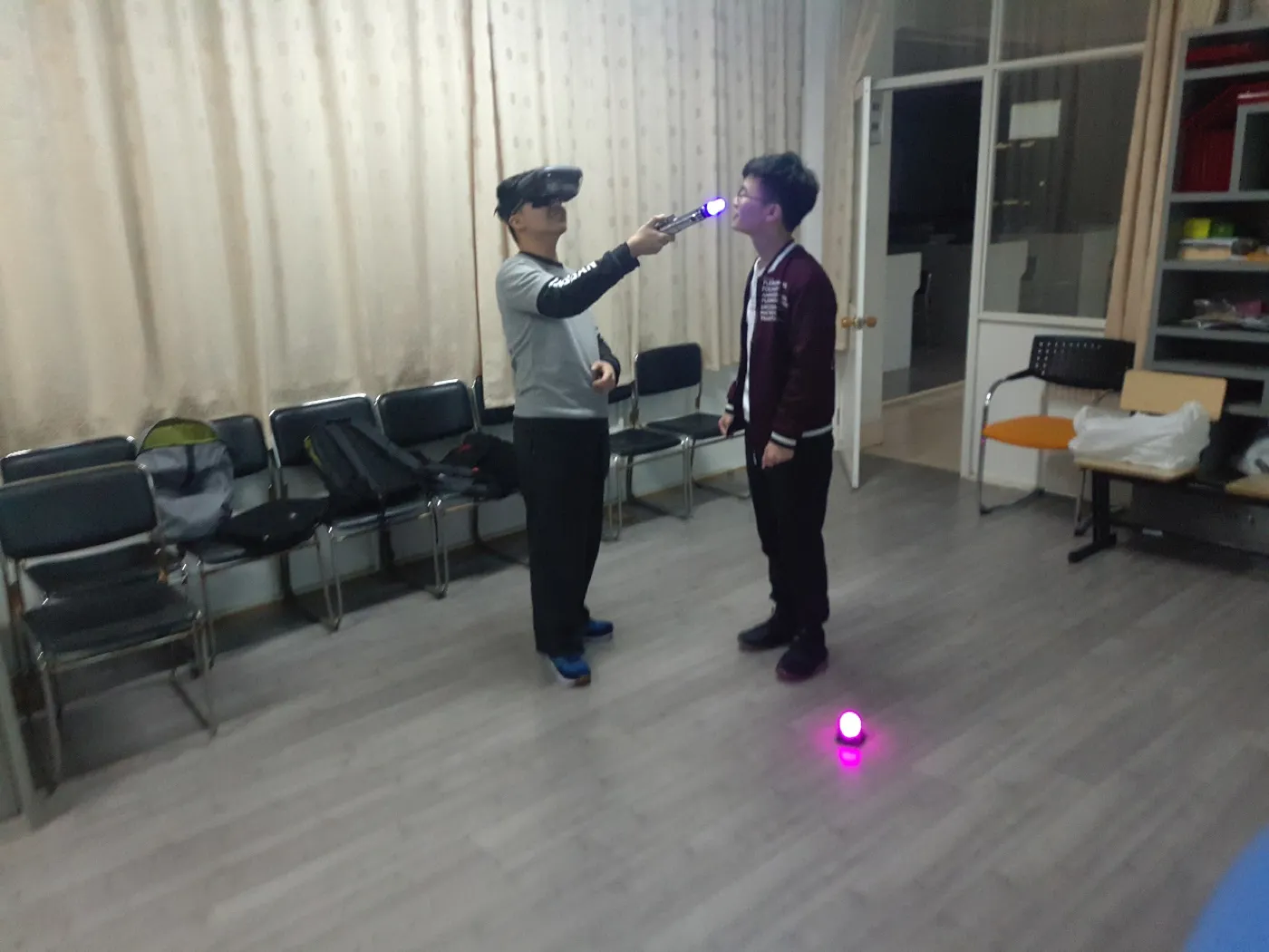 室内空场地上两名学生相对而立,一人戴着头显举起发光的光剑指向对方,另一人侧身看着;地面上有一枚发出紫光的圆形追踪信标,背景是一排排会议椅