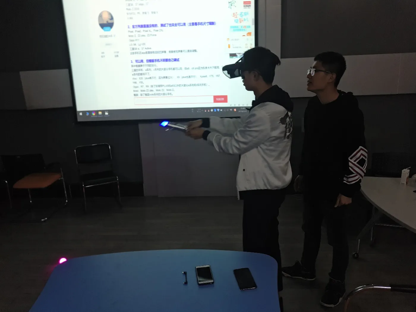 一名学生戴着 Mirage 头显、手持发亮的光剑站立,旁边一人陪着;两人身后的大屏幕上是一个网页,地面有紫色追踪信标,桌上放着两部手机