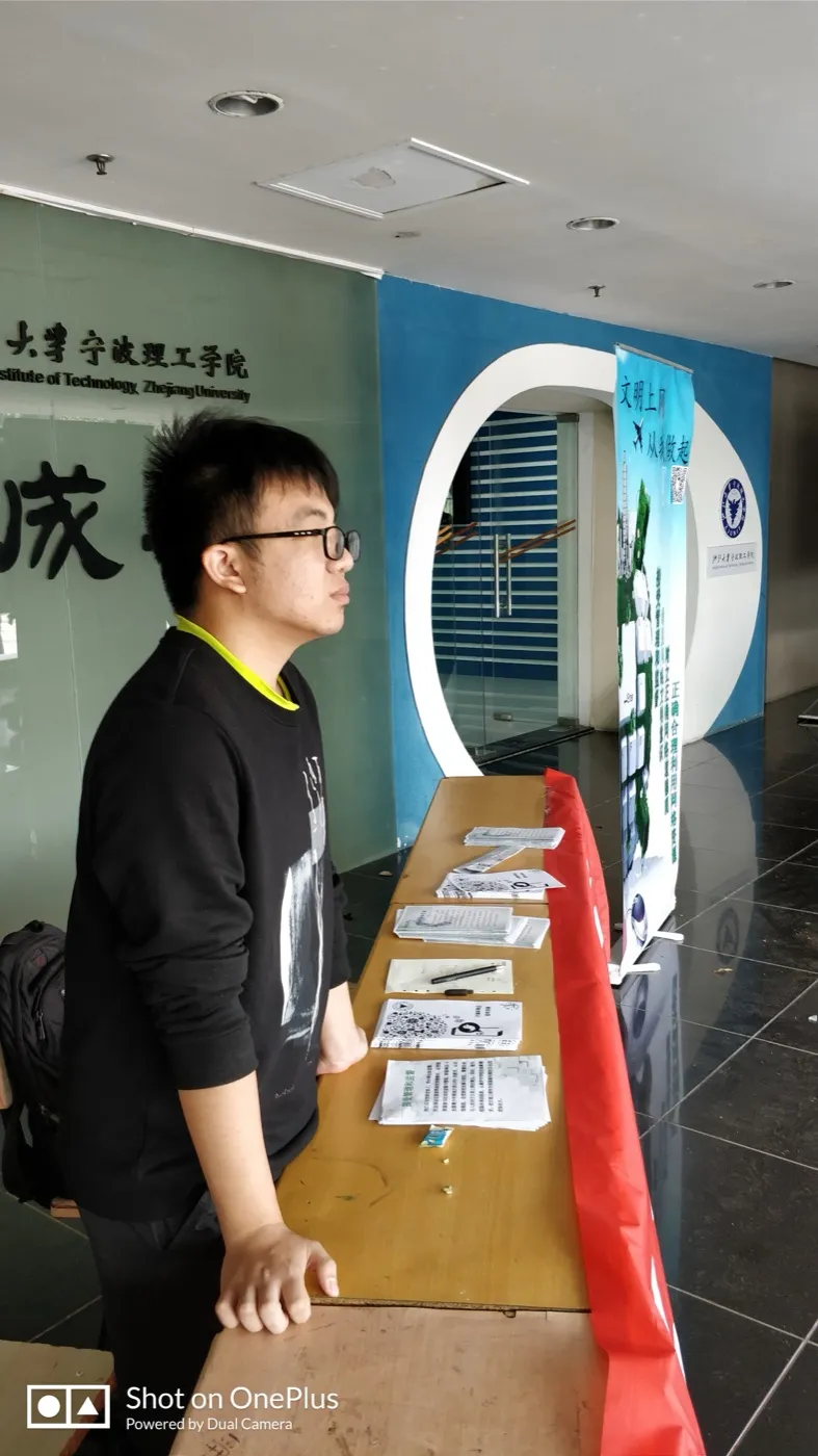 教学楼一楼大厅里，一名戴眼镜的学生站在铺着红布的长条桌旁，桌上分几摞放着宣传单，其中几张印有二维码；身后是一块蓝色易拉宝，上面写着“文明上网 从我做起”与“正确合理利用网络资源”，再往左是刻着“浙江大学宁波理工学院”的玻璃墙
