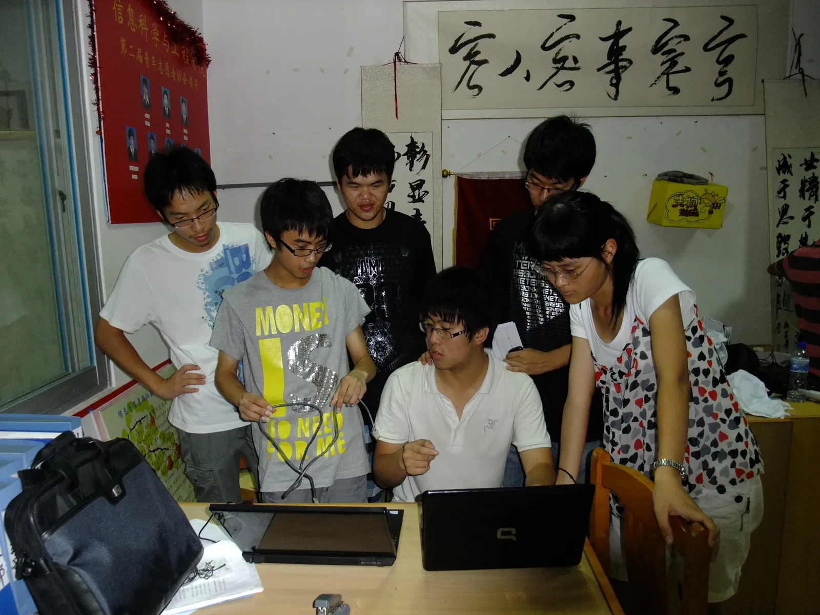 办公室里,一名成员坐在桌前操作笔记本电脑,另外五人站着俯身围看,其中一位手里拎着一根数据线;桌上还摊着一台合上的笔记本与一叠纸。墙上挂着一块贴有成员证件照的红色展板和几幅书法条幅