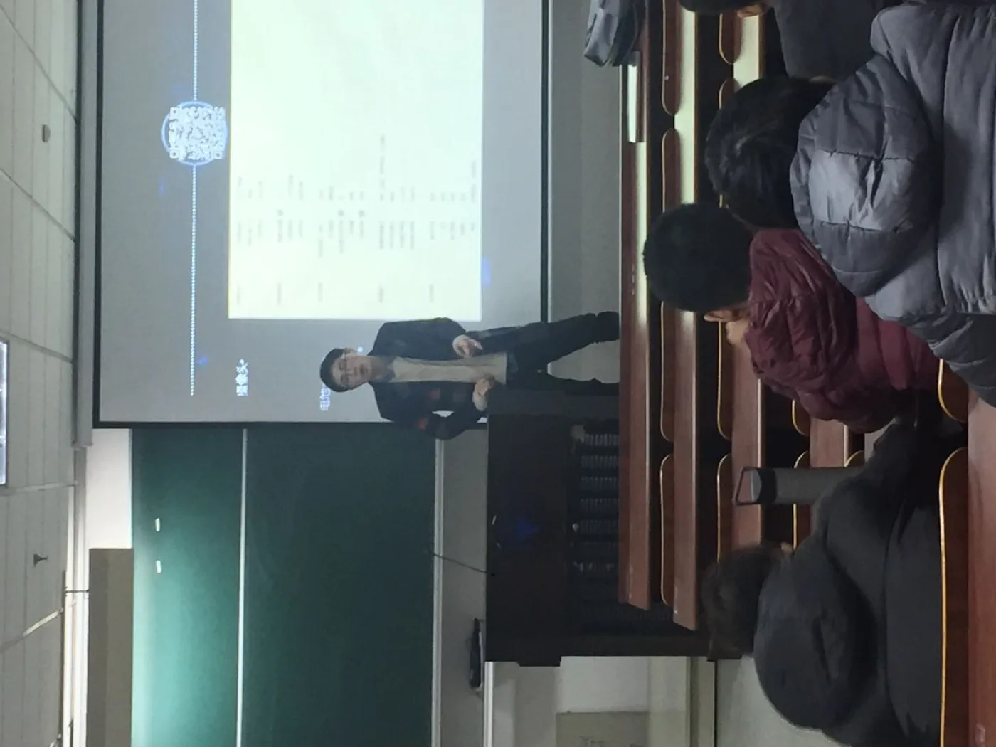 从教室后排隔着几名穿冬装的同学望向讲台,投影幕上是一页浅色幻灯片,一名讲者站在幕布前的讲台边讲解