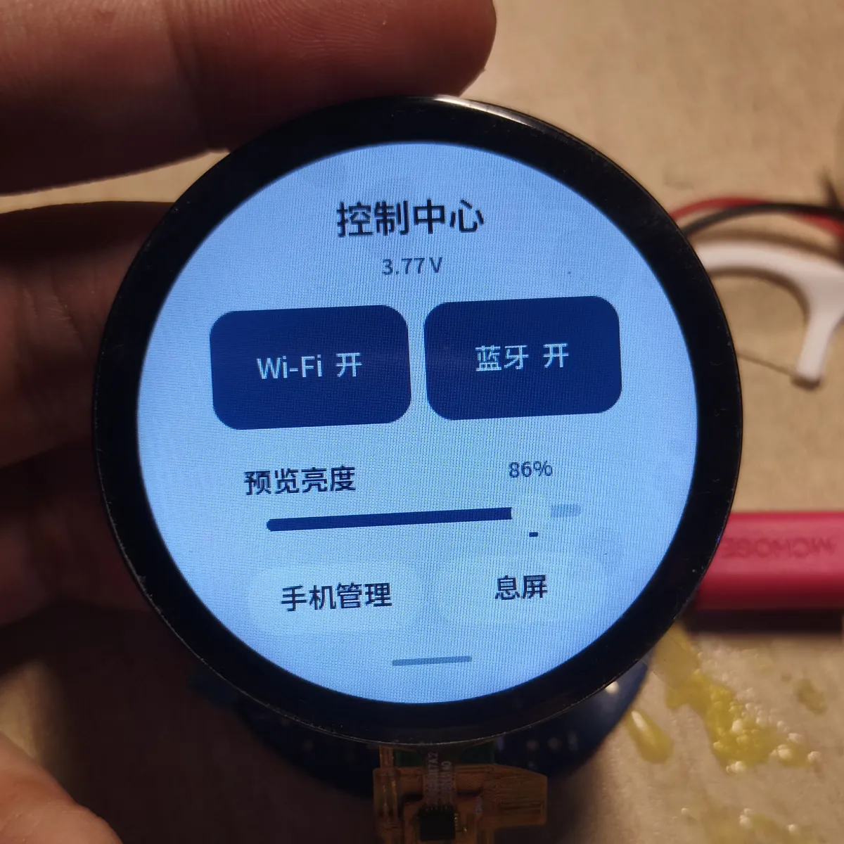手指捏着一块圆形屏幕的电路板,屏幕上是“控制中心”页面:顶部显示电池电压 3.77 V,下面是 Wi-Fi 开、蓝牙开两个按钮、一条预览亮度滑条,以及手机管理和息屏两个按钮