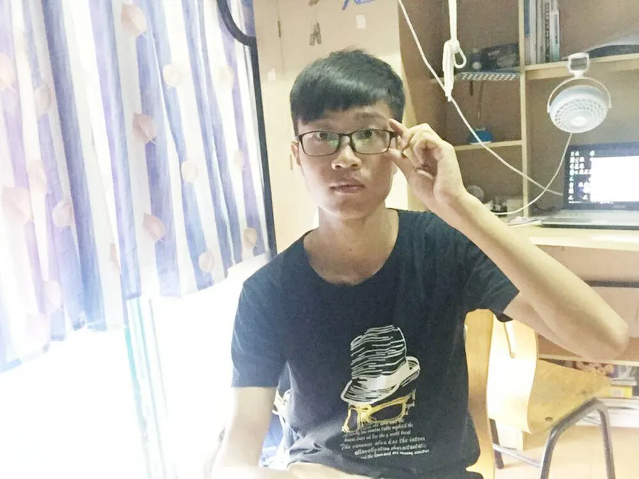 宿舍里的自拍:一名戴眼镜、穿黑色印花 T 恤的男生坐在书桌前,一手扶着眼镜,身后是台灯与书架