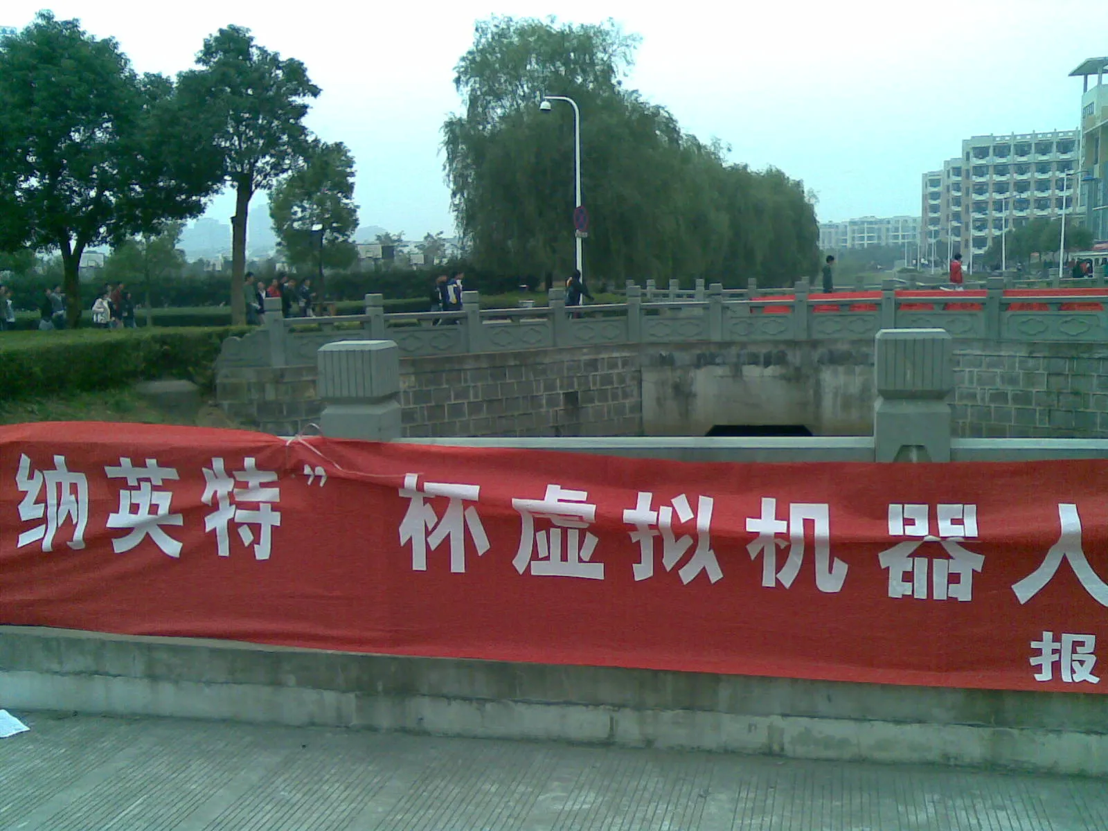 校园河道边的石栏杆下挂着一条红色横幅，白字写着“纳英特”杯虚拟机器人…报…，远处栏杆上还系着另一条红色横幅；背景是柳树、路灯与教学楼