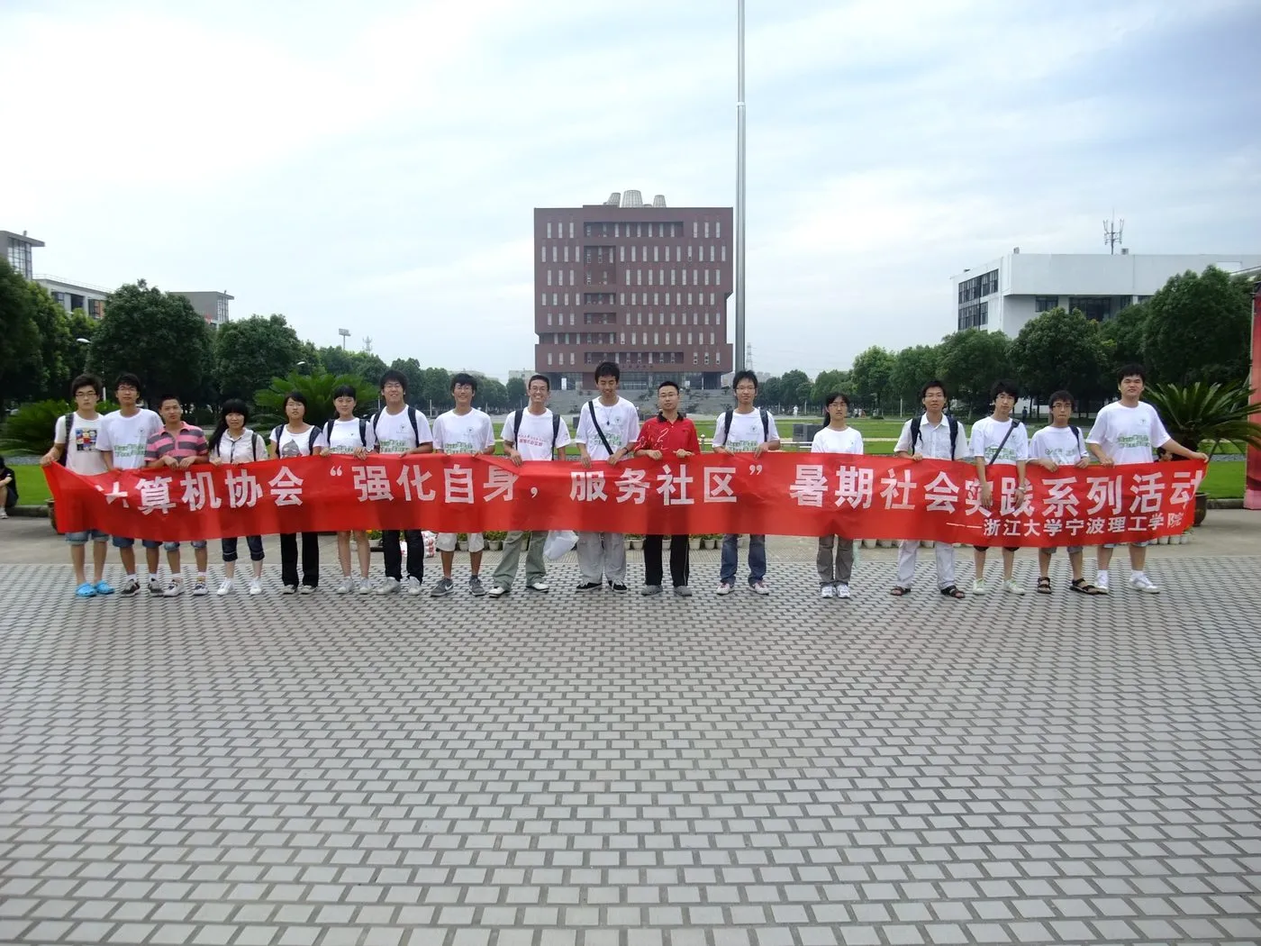 近二十名成员在校园广场上一字排开,共同拉着一条红色横幅,多数人穿着印字的白色队服;身后是旗杆、草坪与一栋红砖高楼。横幅写有“计算机协会‘强化自身,服务社区’暑期社会实践系列活动——浙江大学宁波理工学院”