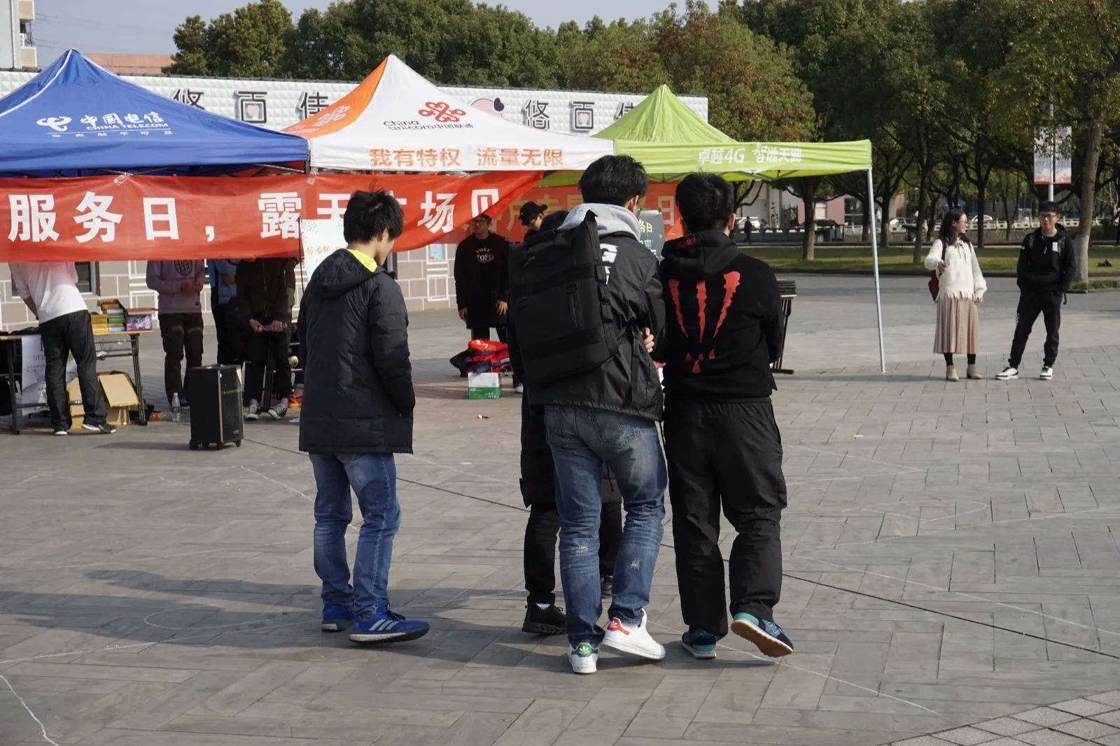 露天广场上支起三顶帐篷,分别印着中国电信、中国联通与一家运营商的 4G 广告;帐篷前拉着一条红色横幅,可见“服务日,露天广场”字样。几名学生背对镜头站在场地中间,远处有人在摊位后整理物品