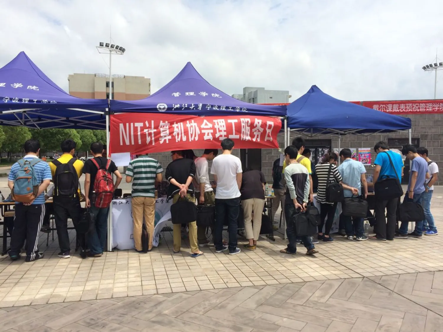 露天广场上并排支着几顶蓝色帐篷,帐篷顶印着“浙江大学宁波理工学院”与“管理学院”;前方拉一条红色横幅,写着“NIT计算机协会理工服务日”。二十来名学生背着包排在长桌前等候,右侧另一顶帐篷的横幅上可见“戴尔课戴表”字样