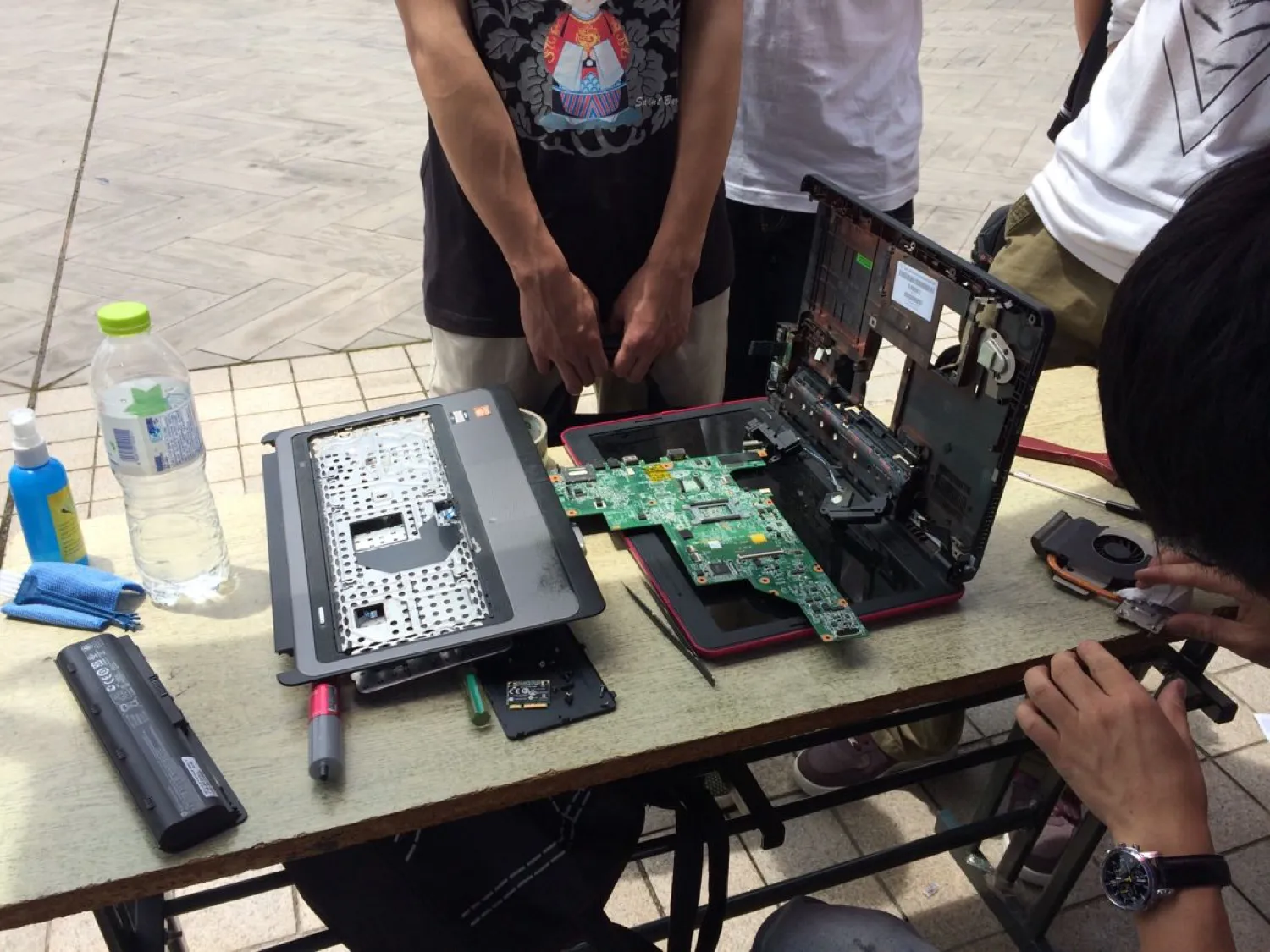 长桌上摊着一台拆到底的笔记本:上盖、卸掉键帽的掌托、单独取出的主板、电池、风扇分开摆着,旁边放着撬棒、清洁喷雾与一块蓝色擦布;两名同学站在桌前看,右侧一只手正在操作
