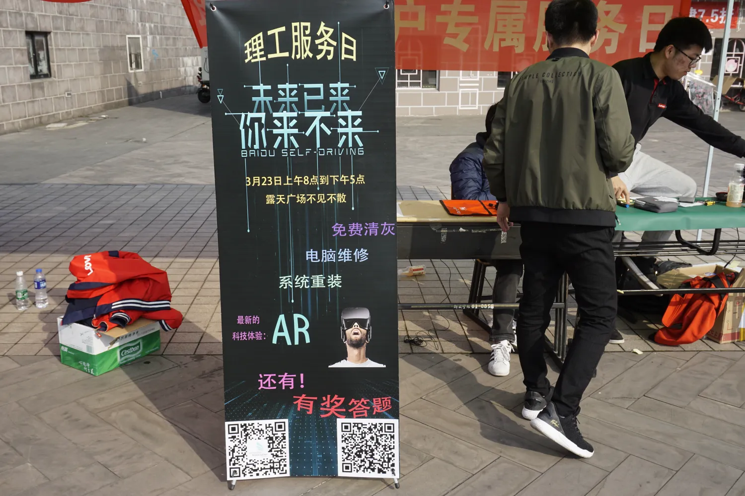 广场地砖上立着一块深色易拉宝,自上而下写着“理工服务日”“未来已来 你来不来”,其下一行英文“BAIDU SELF-DRIVING”,再下是“3月23日上午8点到下午5点/露天广场不见不散”;服务项目列为免费清灰、电脑维修、系统重装,又有“最新的科技体验:AR”并配一张戴 VR 头显的人像,末尾“还有!有奖答题”,底部并排两个二维码。易拉宝旁的纸箱上堆着红色队服,地上放着两瓶水