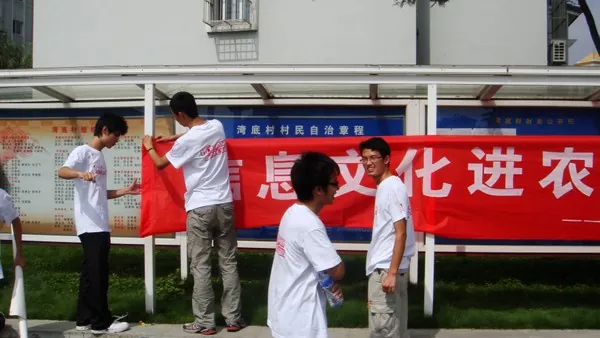 五名穿白色队服的学生在一排村务公开栏前挂一条红色横幅,横幅上“息文化进农”几个大字露在外面;公开栏的蓝色标题写着“湾底村村民自治章程”,背景是村里的楼房