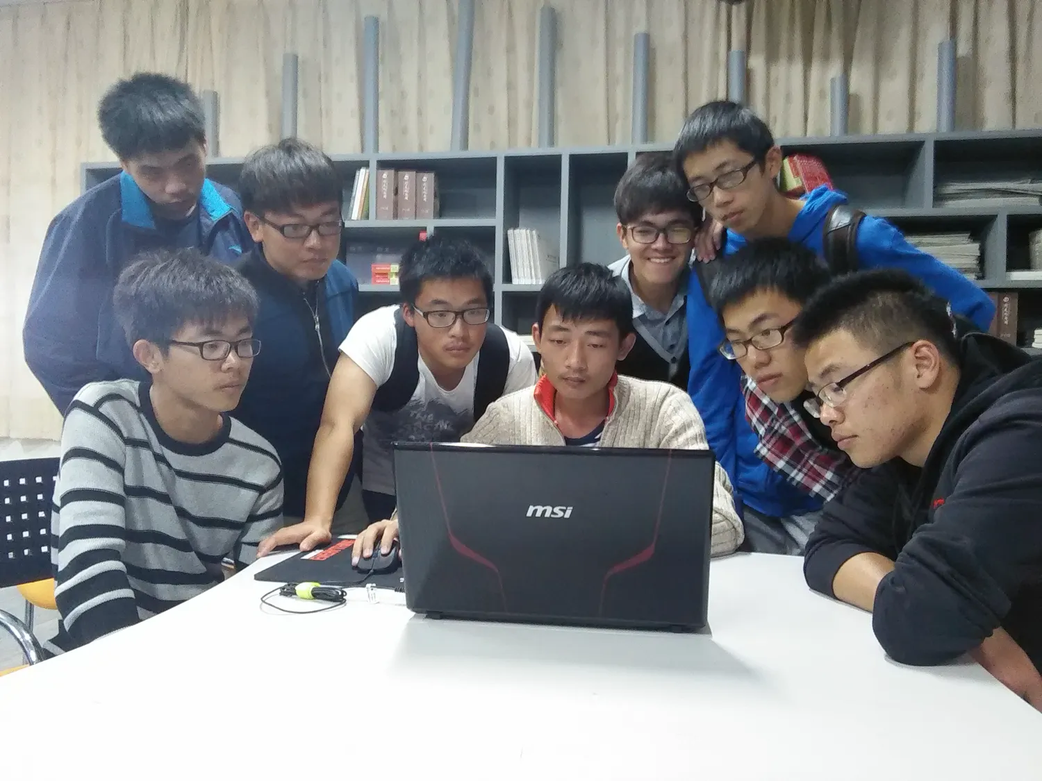 八九名学生紧紧围成一圈,挤在一台 MSI 笔记本前;居中一人手放在触控板上操作,其余人俯身盯着屏幕,背景是书架