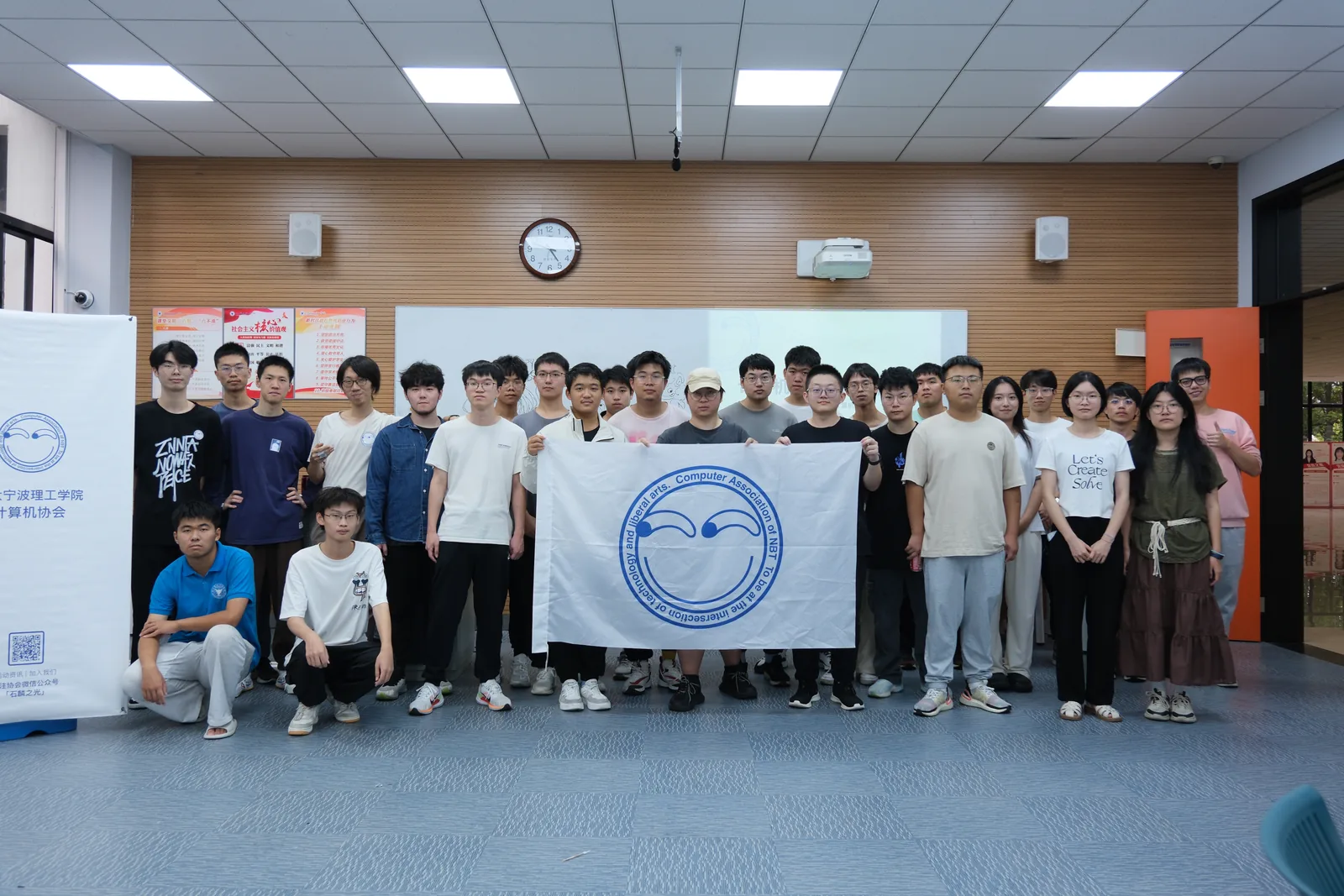 室内合影:二十余人站成两排,多数比出手势或笑着看镜头;左侧立着一块白色易拉宝,上面是协会的圆形笑脸标识