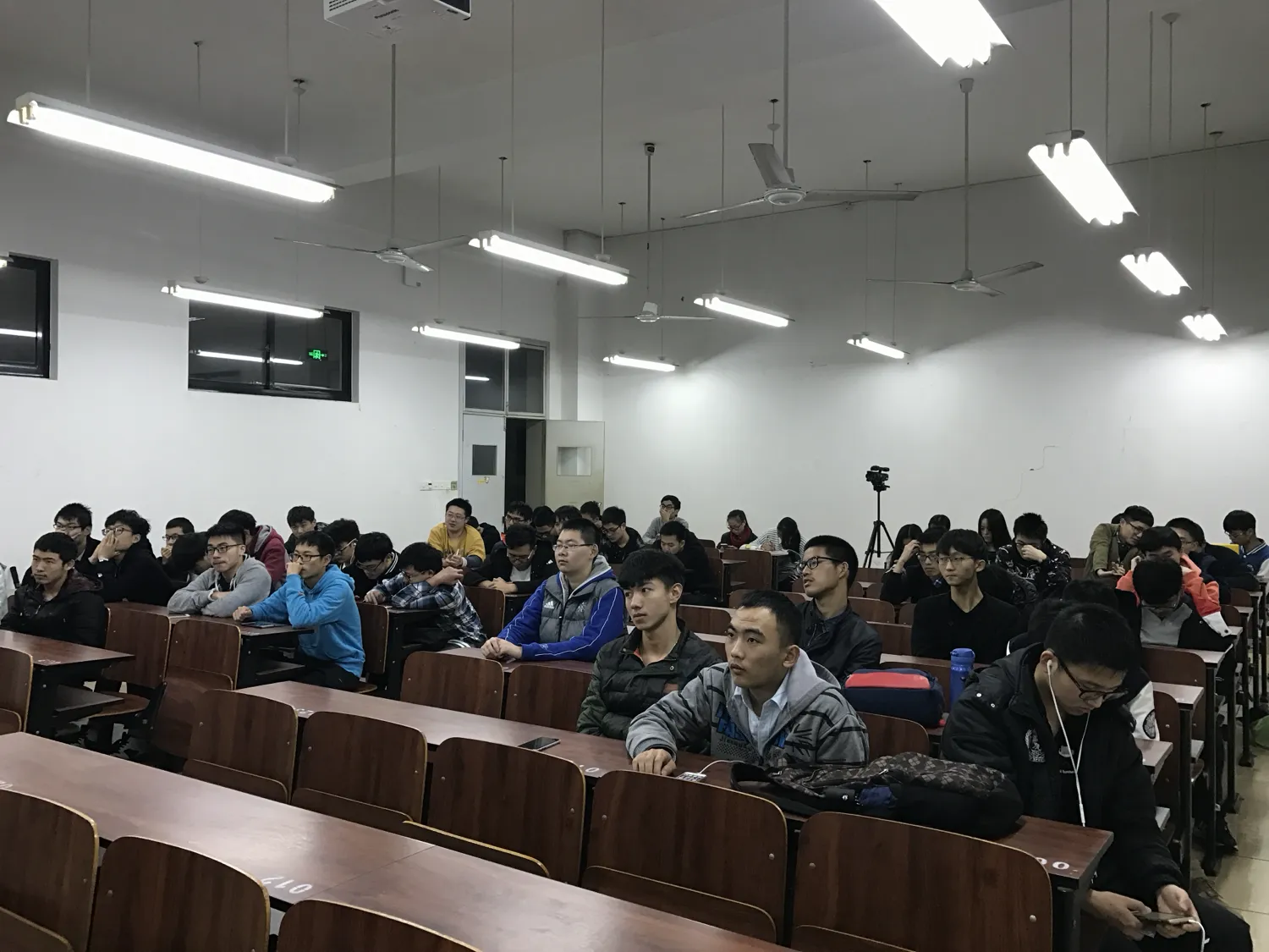 阶梯教室里坐满穿冬装的学生,一直坐到最后一排;靠右过道处架着一台三脚架上的相机,前方讲台亮着