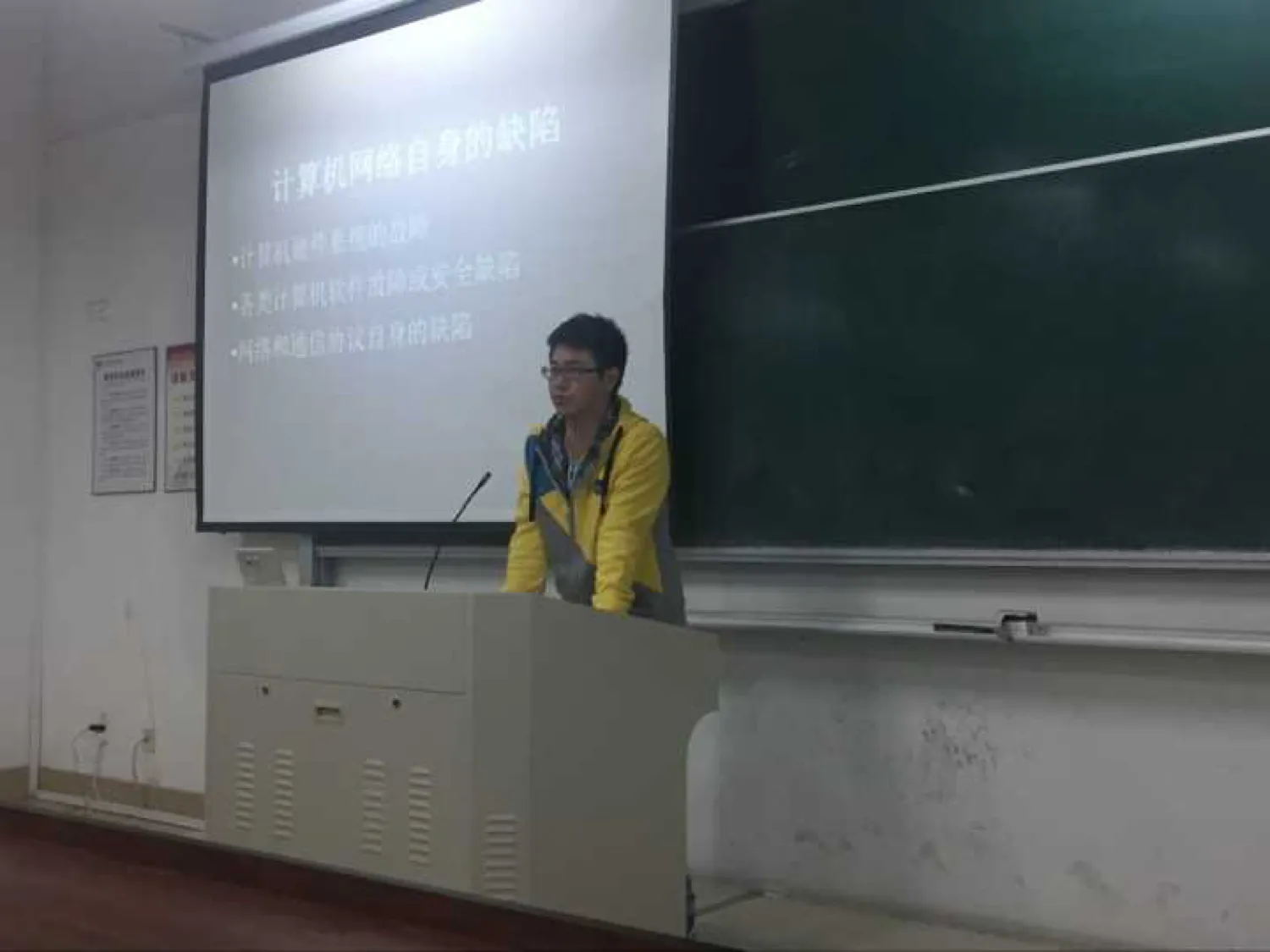 讲台前一名穿黄色外套、戴眼镜的讲者站在话筒后;身后投影幕上是一页标题为“计算机网络自身的缺陷”的幻灯片,下列三条:计算机硬件系统的故障、各类计算机软件故障或安全缺陷、网络和通信协议自身的缺陷